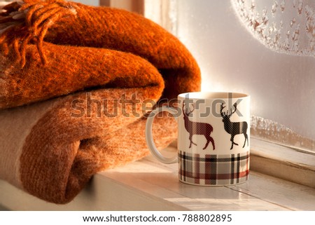 Tea in winter frost on the background. A mug of tea with a picture of deer and a warm blanket on the windowsill. Frost on the glass window. Tea with a warm plaid in winter create coziness.