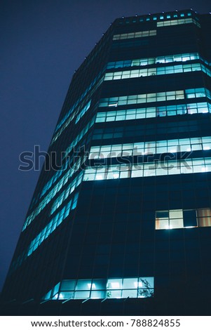 High-rise office building, the night is lit in the windows, people are working. Night view of city skyscraper perspective