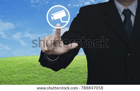 Businessman pressing cctv camera icon over green grass field with blue sky, Business security concept