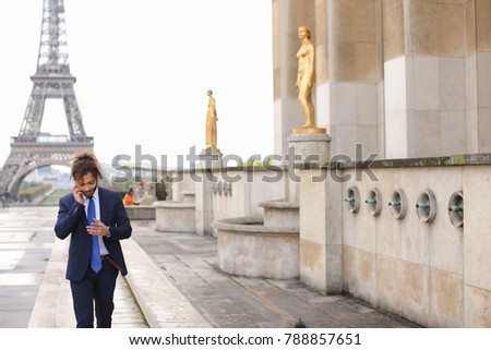 Handsome half Nigerian and French guy calling sweetheart by phone with Eiffel Tower in background. Young man dressed in black suit and blue tie looks cute with curly hair and nice smile. Concept of