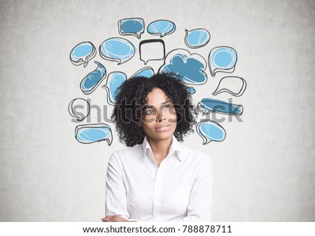 Portrait of a thoughtful African American businesswoman wearing a white blouse and standing with crossed arms. A concrete wall background with blue and white speech bubbles.