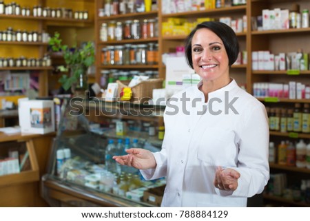 Glad mature female seller in uniform standing with dietary supplements in pharmaceutical  store