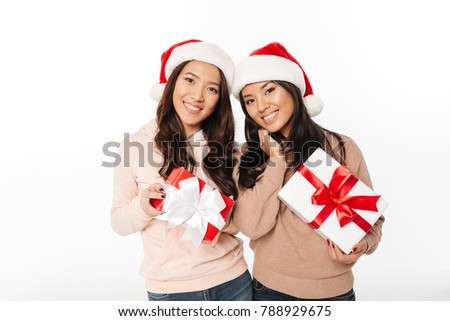 Picture of two asian cute ladies sisters wearing christmas santa hats standing isolated over white background holding gift boxes surprise.