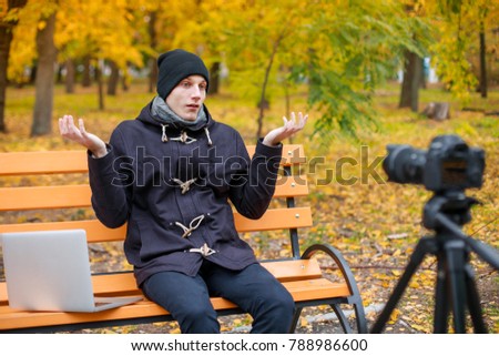 A guy sits in an park on a bench with a laptop in front of a camera on a tripod and disappointed with hands.