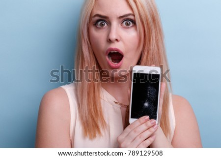 Disappointed girl, holds in hand a broken mobile phone emotionally opening mouth. Blue background.