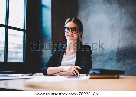 Half length portrait of successful businesswoman dressed in formal elegant jacket smiling at camera.Skilled student in optical eyeglasses sitting at desktop in university interior during break