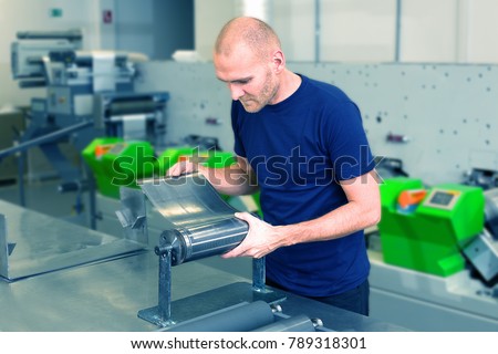 Flexo rotary die cutting preparation. Worker in printing factory, standing next to the printing machine preparing cylindrical die. Operator aligns cutting die on magnetic roll. Label manufacturing. Royalty-Free Stock Photo #789318301