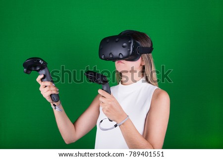 attractive woman wearing virtual reality headset with two handheld trackpads or controllers in green screen studio