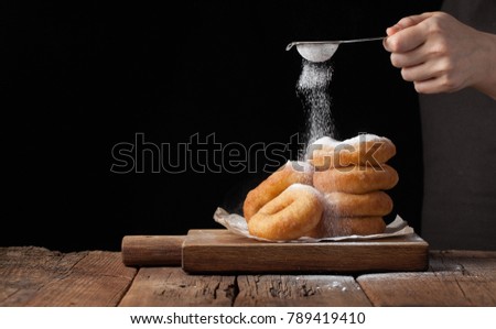 Baker sprinkles sweet donuts with powder sugar on black background. Delicious, but unhealthy food on the old wooden table with copy space