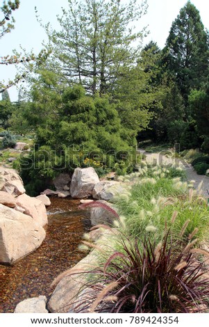 A gentle stream flowing over a rock bed, lined by boulders, Fountain Grass, bushes and evergreens, next to a walking path in Colorado, USA