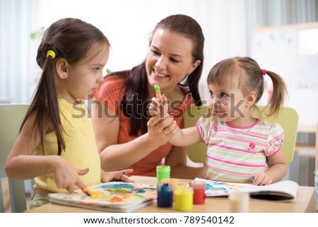 Kids with young teacher woman painting on table together in kindergarten or home.