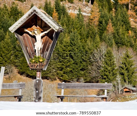 small shrine with image of christ carved in wood with benches typical of mountain trails with forest in the background