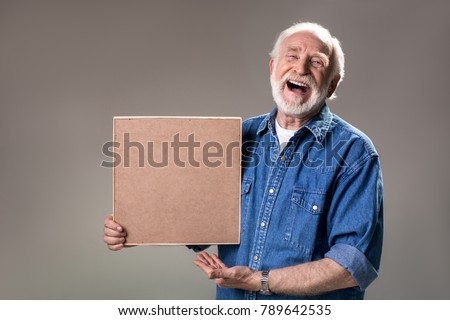 Waist up portrait of happy aging man holding wooden frame, his face expressing unbridled joy. Isolated on grey background