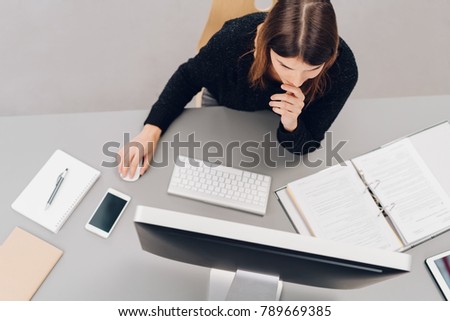 Hard working young businesswoman in the office sitting reading a desktop monitor with a thoughtful expression in an overhead view