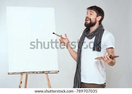 man with a brush and a palette stands at the easel with a canvas on a light background                              