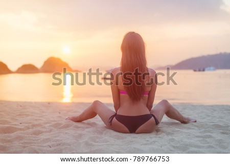 Back view of young female with long brown hair sitting on beach legs spread far apart in black bikini panties admiring sunset and seascape.
