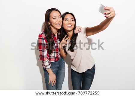 Picture of two asian cheerful positive ladies sisters. Looking aside make selfie by mobile phone.