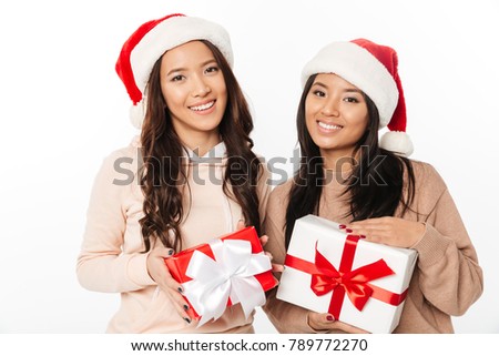 Picture of two asian cute ladies sisters wearing christmas santa hats standing isolated over white background holding gift boxes surprise.