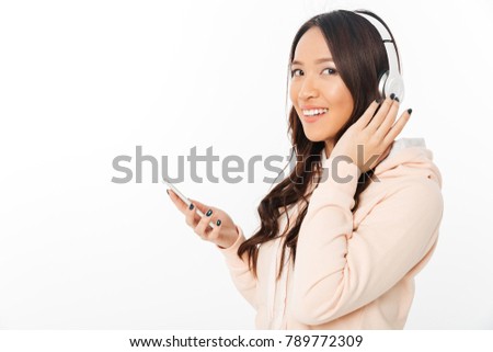 Picture of asian happy woman standing isolated over white background listening music with headphones using mobile phone.