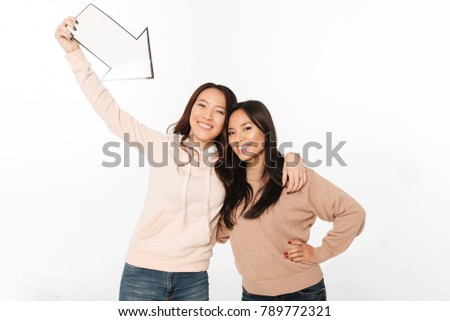 Photo of two asian positive ladies sisters standing isolated over white background holding arrow.