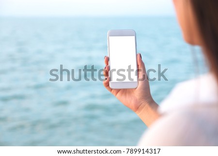 Mockup image of a woman's hand holding white mobile phone with blank desktop screen by the sea and blue sky background