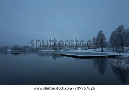 Beautiful nature and landscape photo of snowy blue dusk evening at beach in Katrineholm Sweden Scandinavia. Nice cold winter day with soft light and sky. Lake, trees and wooden bridge. Calm, peaceful.