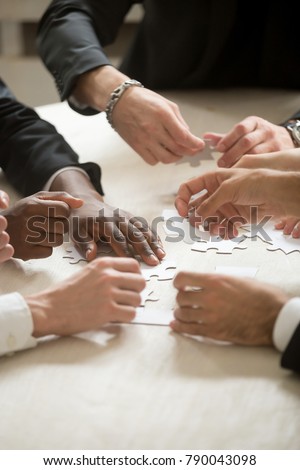 Multi-ethnic group of people assembling jigsaw puzzle together, hands joining pieces at desk, successful teamwork concept, team building activity, help and support in business, close up vertical view