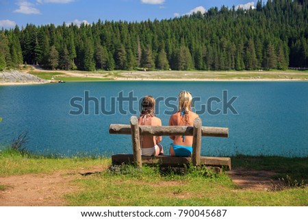 back view of two girls in bikini sitting on a bench near a mountain lake and pine trees. Black lake, Montenegro