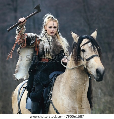 Beautiful furious scandinavian viking warrior woman with war makeup in traditional clothes riding a horse in a forest with ax in hand, threatening. Lagertha look. Series
