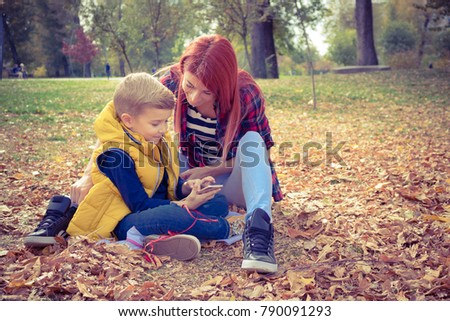 Happy boy and his mother relaxing on autumn leaves and watching something on mobile phone together.