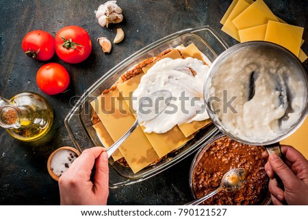Woman cooking homemade classic lasagna bolognese, on dark blue table; with ingredients, top view copy space, hands in picture