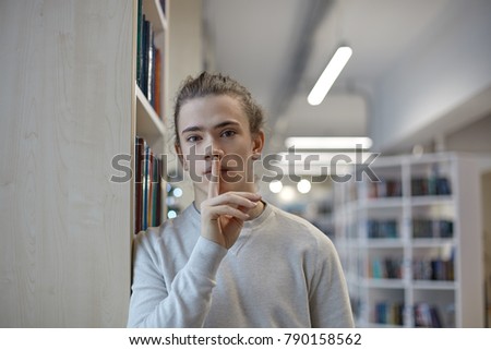 Portrait of serious high school student in casual clothes standing in library and showing silence gesture, asking to keep silence and not make noise because he is studying, preparing for exam or test