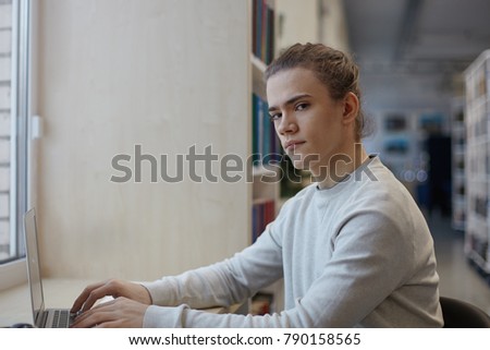 People, modern lifestyle, technology and elecronic gadgets concept. Portrait of positive stylish young European man sitting at window at cafe and typing something on laptop pc, looking at camera