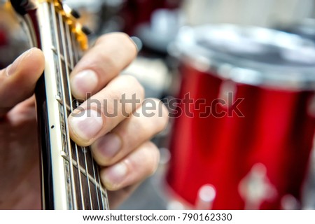 The guitarist's hand, close-up and soft focus, takes the akrod on a guitar fretboard, against the background of the drum set