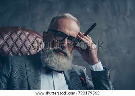 Close up of dreamy gentlemen,  selective, bearded investor in glasses, hold hand with cigar on temple, nicotine, habit, in tuxedo with bow tie, jacket, sit in leather armchair over gray background