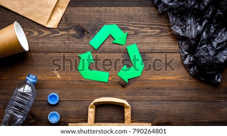 Green paper recycling sign among waste materials paper, plastic, polyethylene on dark wooden background top view copy space