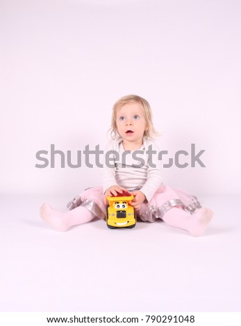 Little blonde toddler girl with big Blue Eyes in pink fluffy skirt  play with yellow toy. Sitting on the floor, white background. Portrait, close-up isolated 
