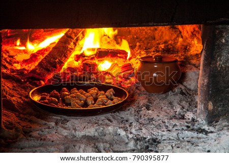 Clay pot and frying pan with meat stands in a Russian oven on an open fire on wood, stewed roast. Rustic authentic food.
