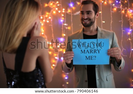 Young man holding paper with text WILL YOU MARRY ME? in front of his girlfriend on engagement day