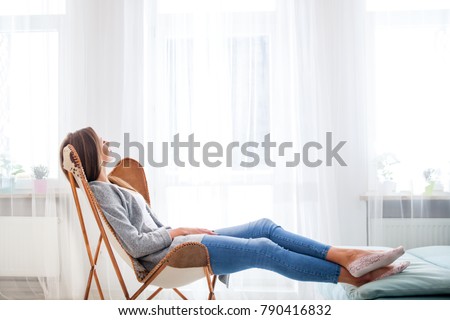 Young woman at home sitting on modern chair near window relaxing in living room Royalty-Free Stock Photo #790416832