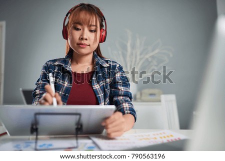 Concentrated young photo editor listening to music in headphones while using graphics tablet, interior of modern office on background