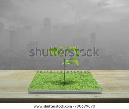Small tree growing from an open book on wooden table over pollution city tower, Ecological concept