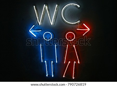 Close up glowing blue and red neon lights of WC toilet sign with directions to men and women, left and right arrows, over dark black wall background