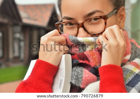 Portrait of an Asian girl in glasses, she screwed her face with a scarf, only her eyes and hands are visible. The hair is tied to the knot. Standing on the street. Bright colours. Horizontal photo