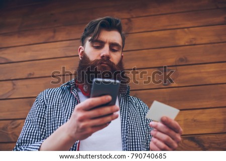 Below view of bearded hipster guy with business card in hand dialing telephone number to call on digital smartphone device standing on wooden promotional background for your advertising message