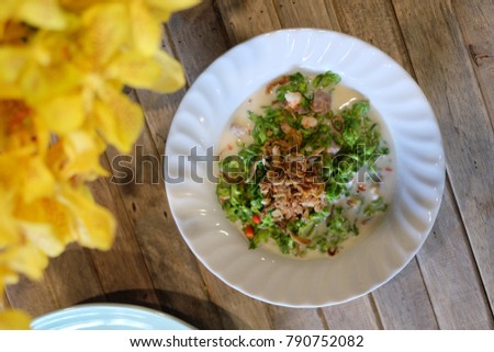 Traditional Thai-style Spicy stir-fried cowslip creeper salad with coconut milk, chilies, and fried shallots all in a white  plate by a vase of yellow orchid and on wooden plank table. Top view pic.