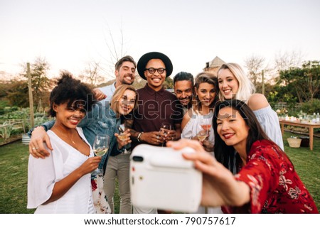 Woman with friends taking selfie using instant camera. Group selfie at outdoor party.
