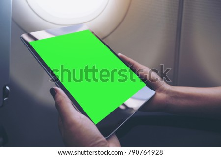 Mockup image of a woman holding and looking at black tablet pc with blank green desktop screen next to an airplane window with clouds and sky background