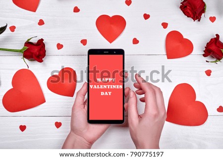 Close up of mobile smart phone with sign Happy valentines day in woman hand, red hearts and flowers on wooden background, copy space. Flat lay, top view. Valentines day application mock up template