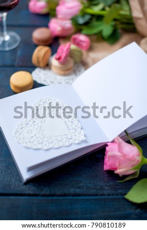 A bouquet of roses is pink, in gray paper and a white heart. Open notebook. A glass of red wine. Sweet pasta macaroons of different colors, on Valentine's Day. A free place for text or advertising.
